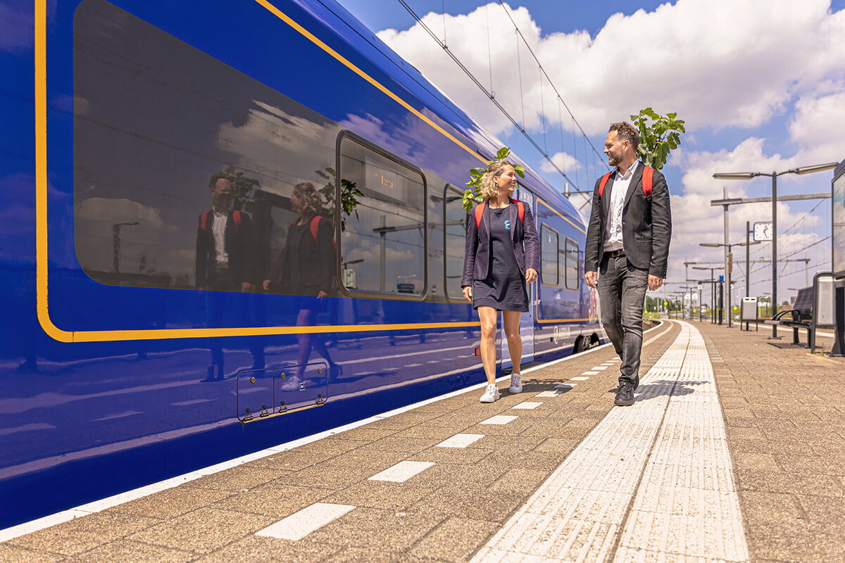 Beeld van de Campagne elke beweging telt:  Twee mensen op perron bij trein met boompje in een rode rugzak. Ze kijken naar elkaar terwijl ze op het perron lopen. Naast ze staat een blauw Arriva trein stil.