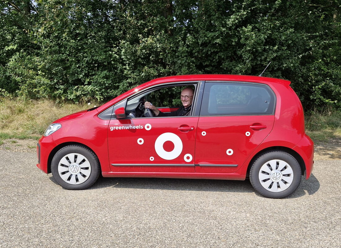 Rode Greenwheels deelauto staat stil op een parkeerplek. In de auto zit wethouder Jan Weling. Op de achtergrond staan groene struiken 