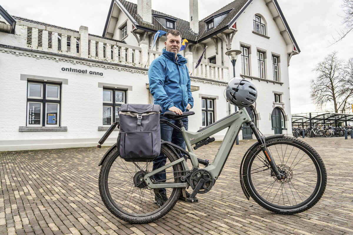 Jean Paul Urlings in blauwe jas met een speedpedelec voor het witte station van Meerssen