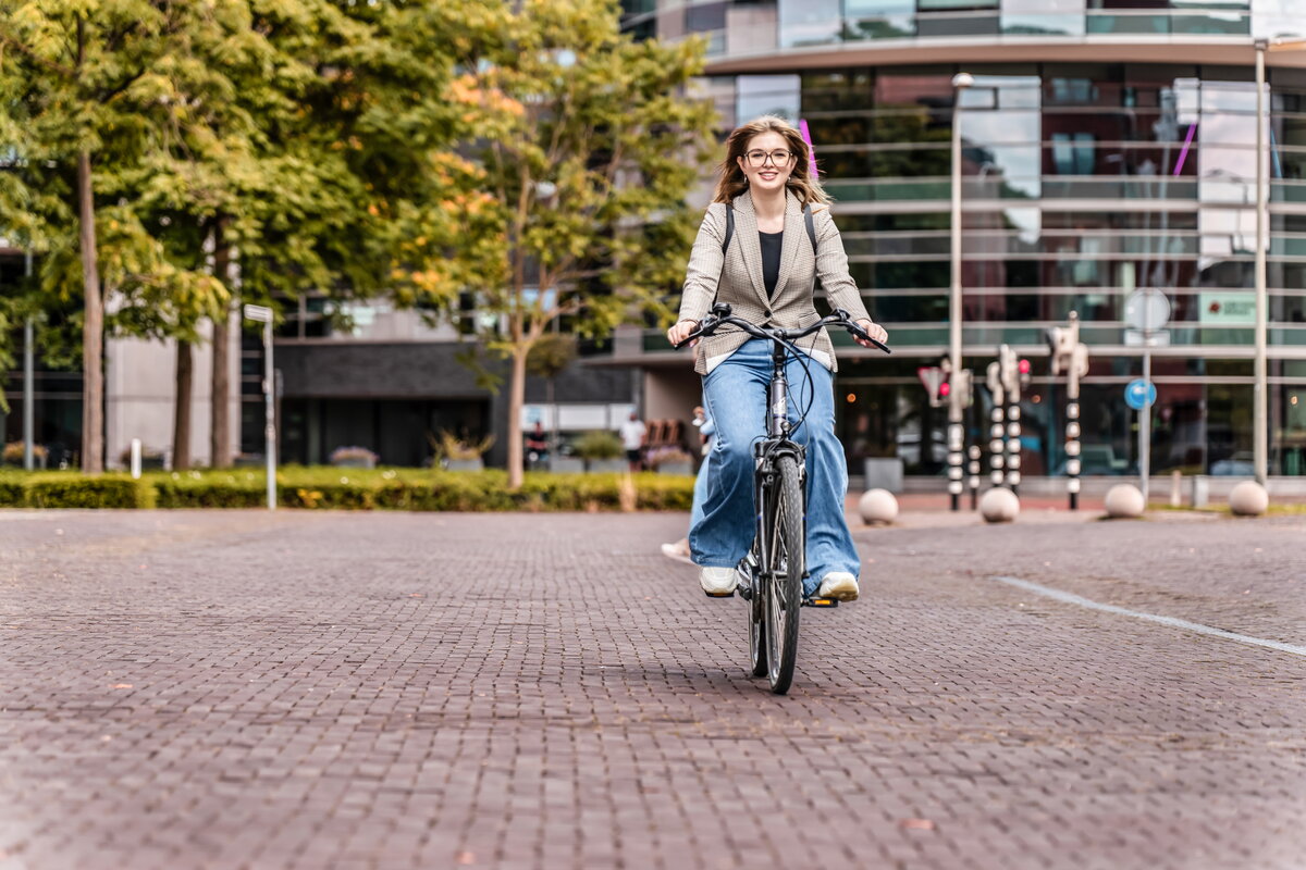 Jonge vrouw op de zwarte fiets, fietst richting de camera in stedelijke omgeving in het voorjaar. Op de achtergrond zie je gebouwen van Maastricht, bomen en verkeerslichten. Meisje heeft bruine lange haren, draagt een bril, bruine blazer en blauwe jeansbroek.