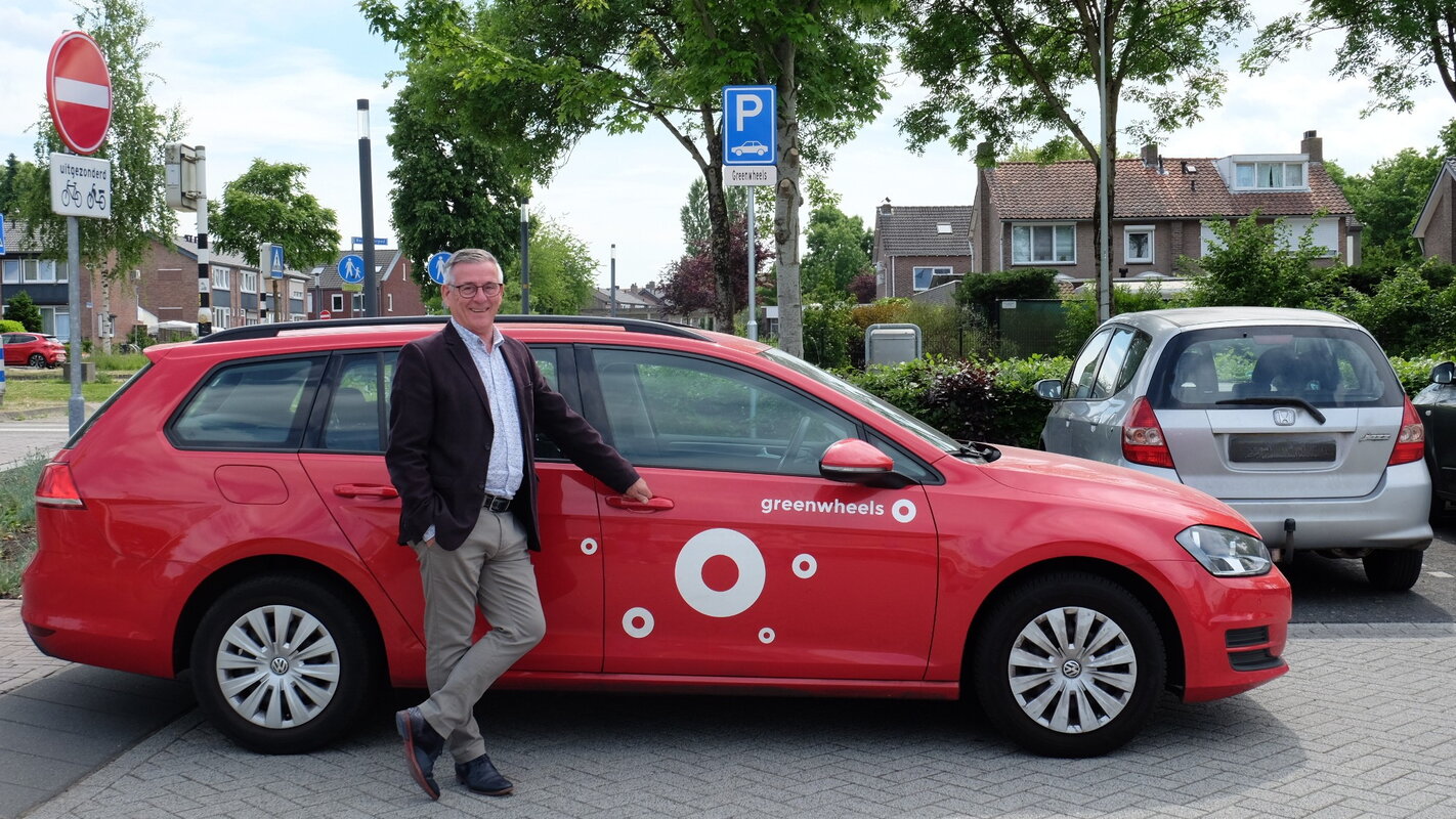 Rode Greenwheels deelauto in gemeente Landgraaf met wethouder Janssen. Wethouder Janssen, een man, leent tegen de rode Volwswagen deelauto van Greenwheels die stil staat op een parkeerplaats