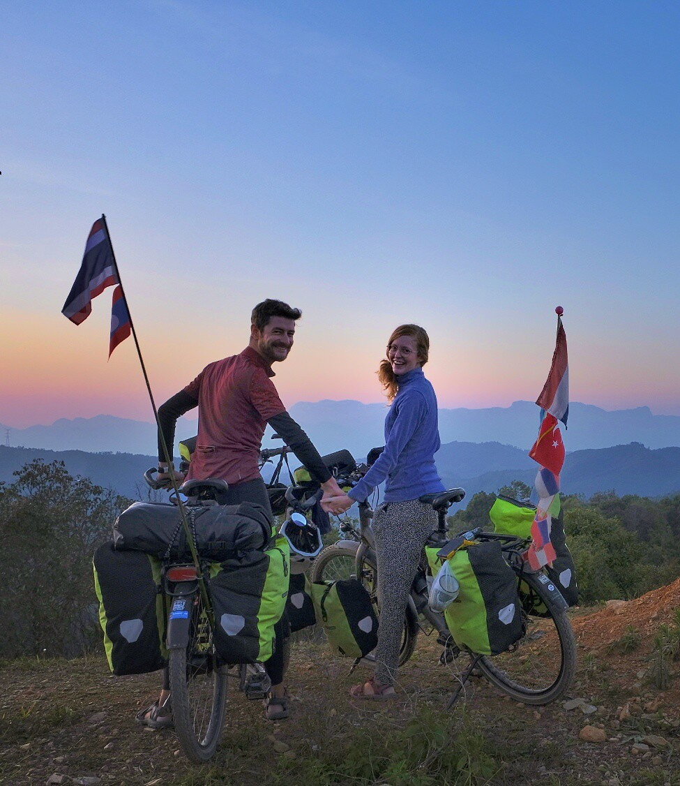 Eloy en Audrey op de fiets met vlaggen van verschillende landen. In de verte zie je bergen