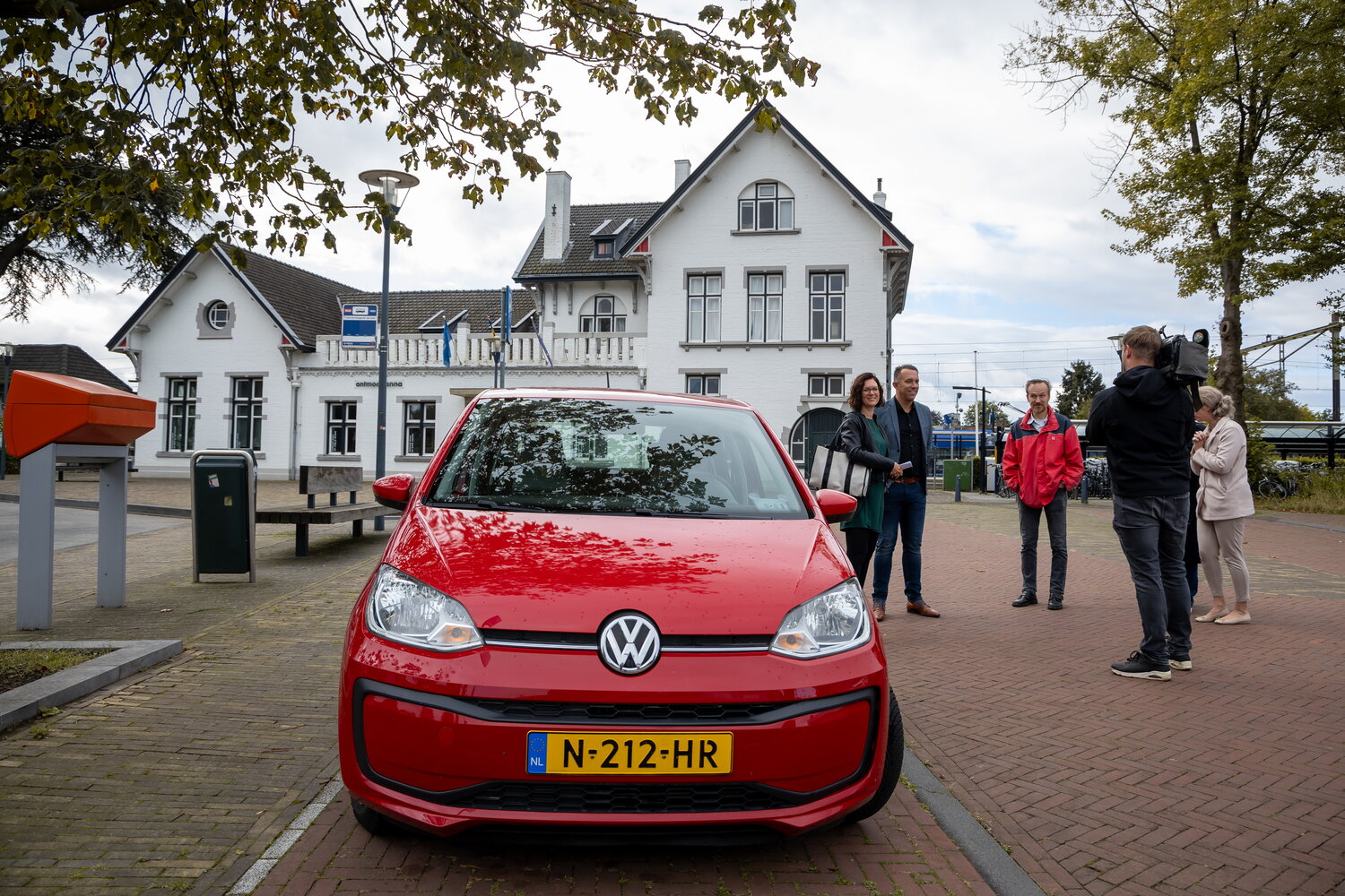 Een rode Greenwheels deelauto van Volkswagen met daarnaast een groep mensen en cameraploeg. Op de achtergrond het witte station van Meerssen 