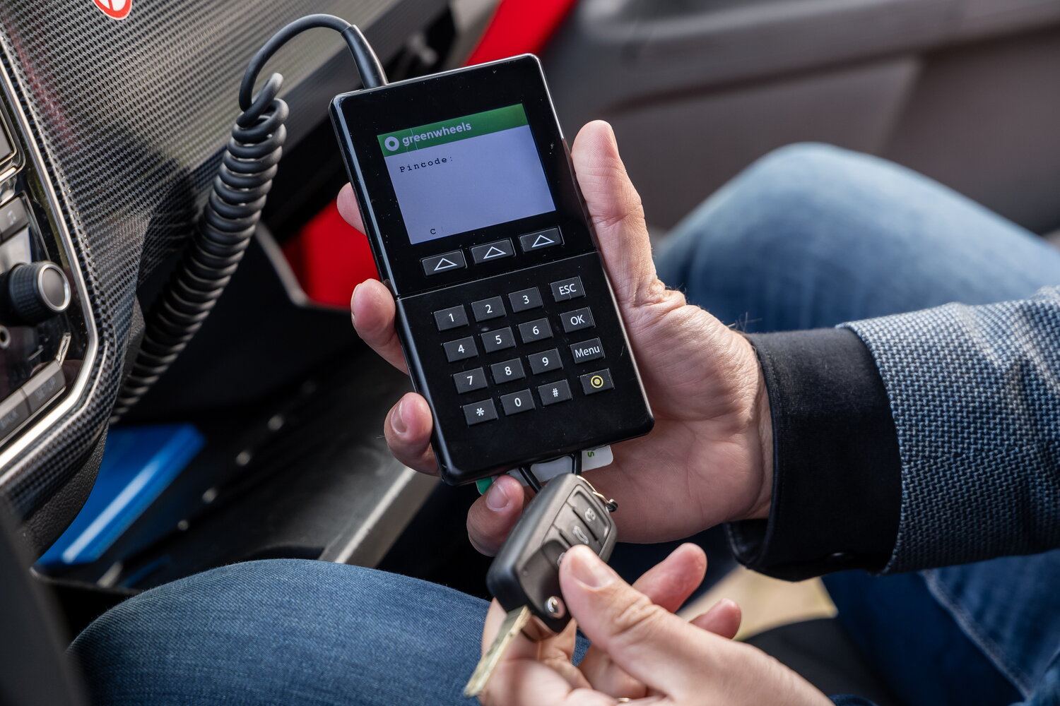 Een mensenhand houdt een apparaat vast waarmee je de app van Greenwheels gebruikt en een autosleutel
