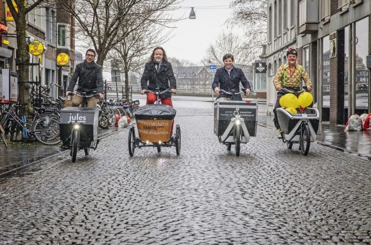 Vier bakfietsen rijden naast elkaar in Maastricht. In een bakfiets liggen gele ballonnen.
