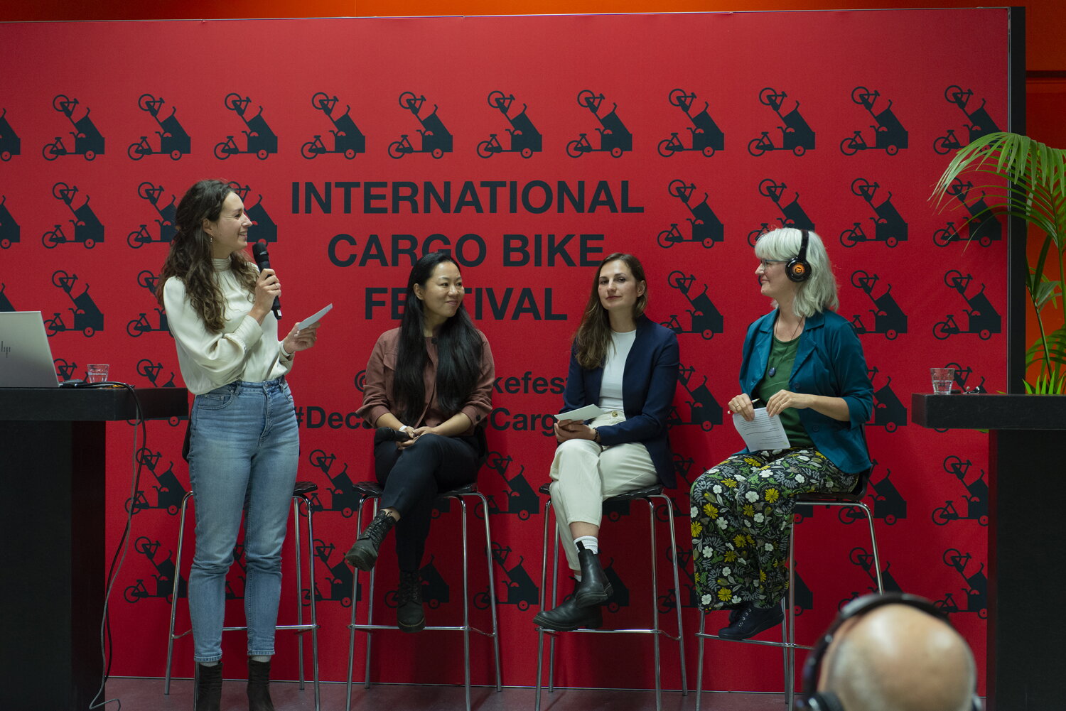 Vier vrouwen zijn in gesprek tijdens het Cargobikefestival. Op de achtergrond een rode wand met het logo.