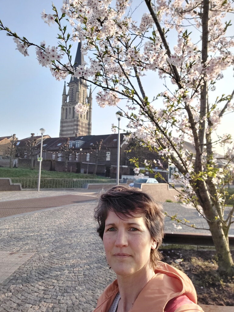 Johanneke Koelstra wandelend in Sittard naar haar werk bij de gemeente. Johanneke maakt een selfie met op de achtergrond de kerk van Sittard, woonhuizen en een bloesemboom